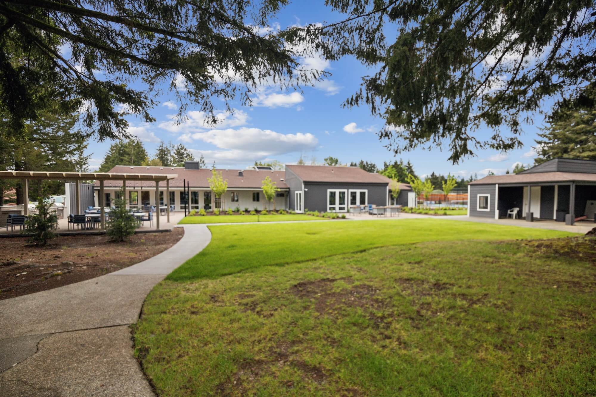 Outdoor community area with green lawns, pathways, covered seating areas, and surrounding buildings under a partly cloudy sky in Olympia